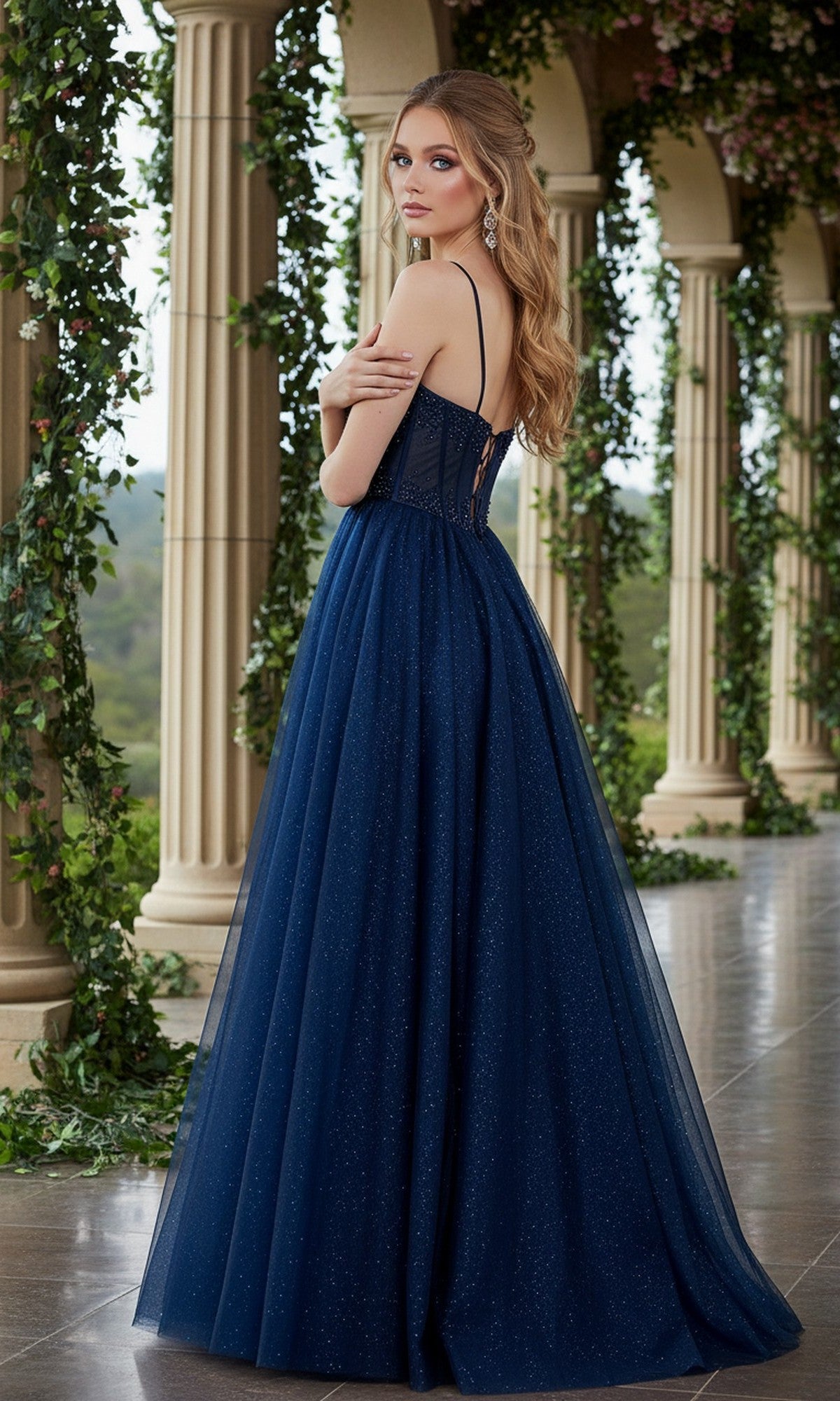 Wearing the Long Prom Dress: Cinderella Couture 8459J, a woman in a navy blue glitter princess gown with an a-line tulle skirt stands on a columned terrace with greenery, looking over her shoulder.